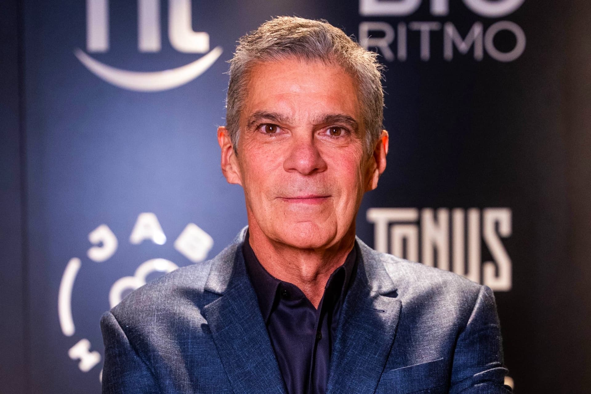 Foto de Edgard Corona, homem com cabelo grisalho, curto, com terno azul e posando em frente a um painel com um logo de suas empresas.